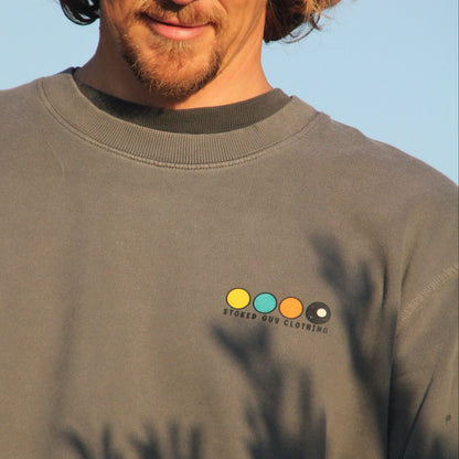 A man wearing a beige baseball cap and a dark gray crewneck sweatshirt with a small colorful logo on the left chest, photographed outdoors against a blue sky.