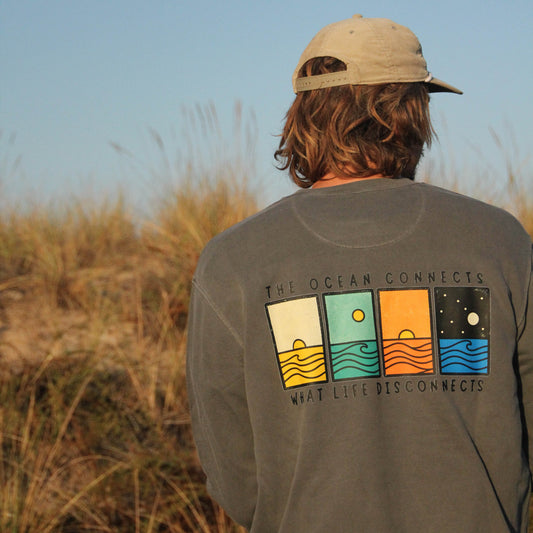 A person viewed from behind wearing a beige baseball cap and a dark gray long-sleeve t-shirt with a colorful graphic on the back. The background shows dry beach grass under a clear blue sky.