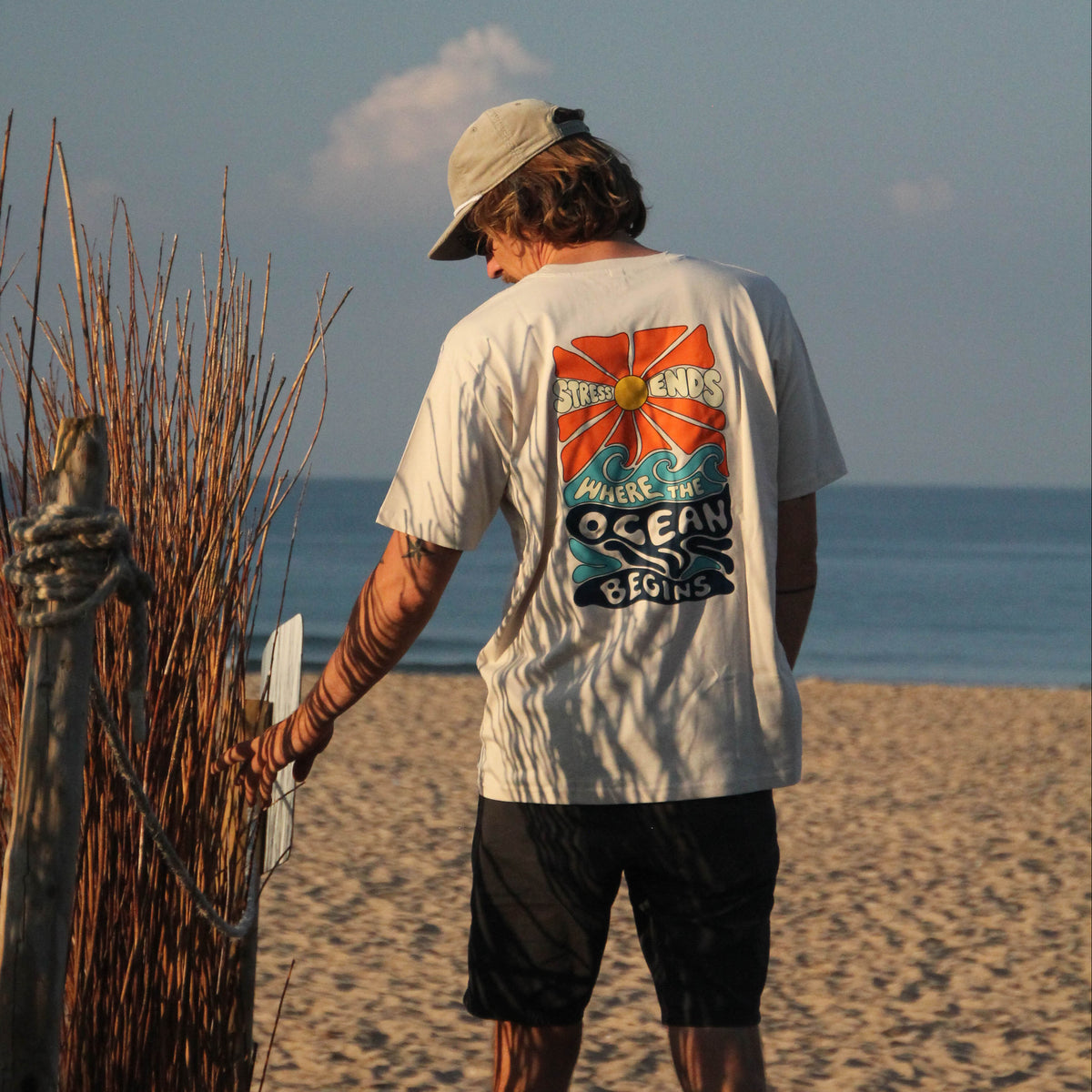 A man standing on a sandy beach facing the ocean, wearing a white graphic t-shirt with a colorful 'Ocean' print on the back, black shorts, and a beige cap.