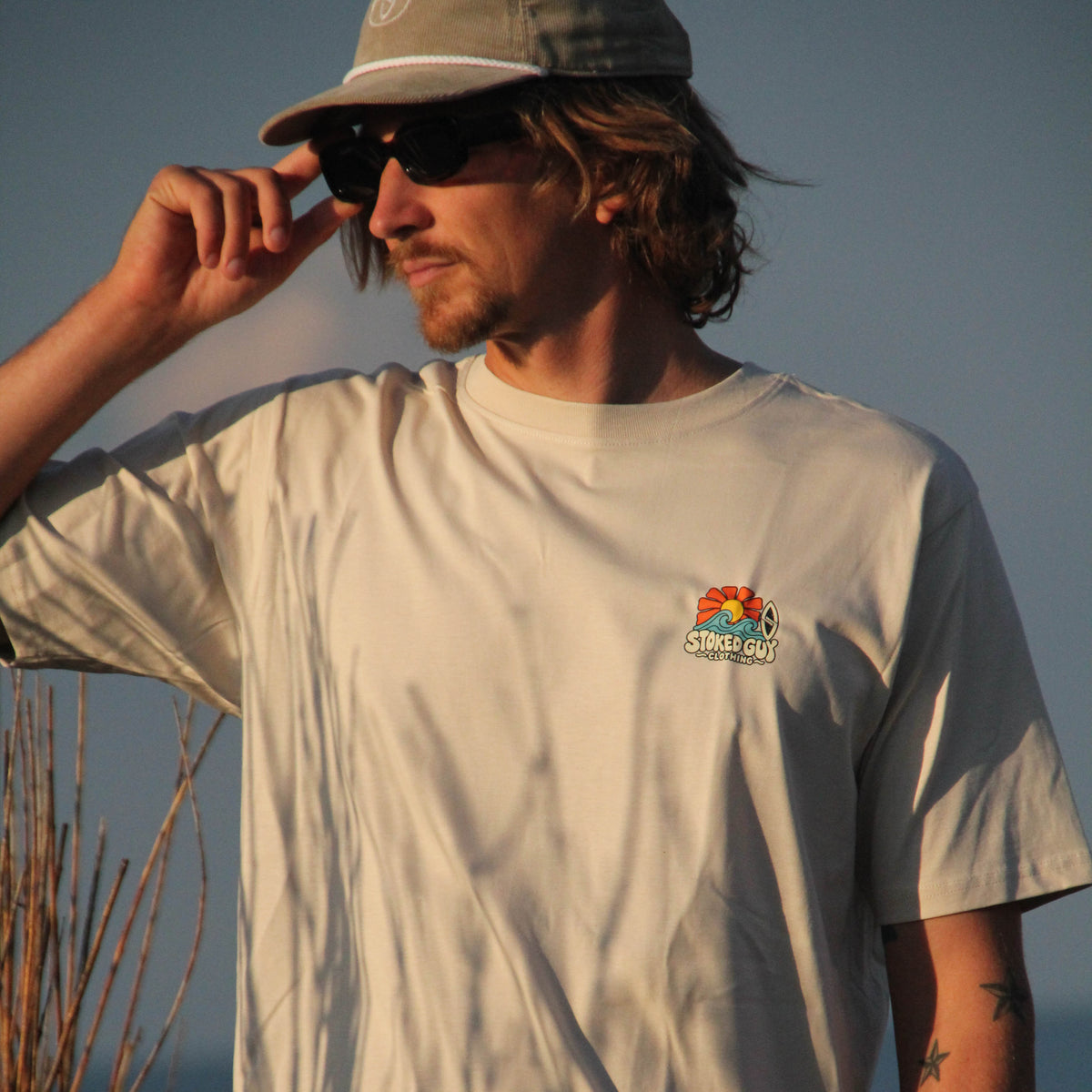 A man wearing a light beige short-sleeve t-shirt with a small colorful 'SUNSHINE' graphic on the left chest, a gray cap, and sunglasses, standing outdoors near a beach with warm golden hour lighting.