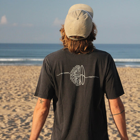 A man standing on a sandy beach with the ocean and blue sky in the background, viewed from behind. He is wearing a beige baseball cap and a dark charcoal gray t-shirt with a white graphic print on the back.
