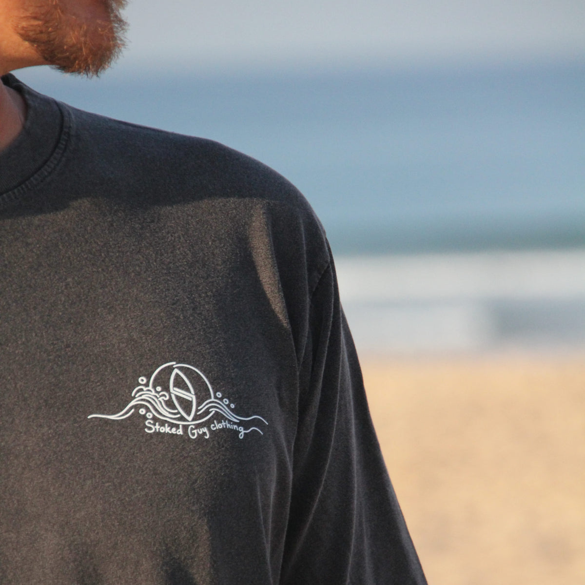 A close-up of a man wearing a faded black t-shirt with a white graphic on the left chest, set against a blurred beach background.