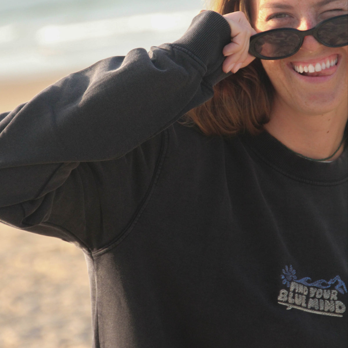 A woman smiling on a beach while wearing a black crewneck sweatshirt with a blue graphic logo that reads 'Find Your Blue Mind' and black sunglasses.