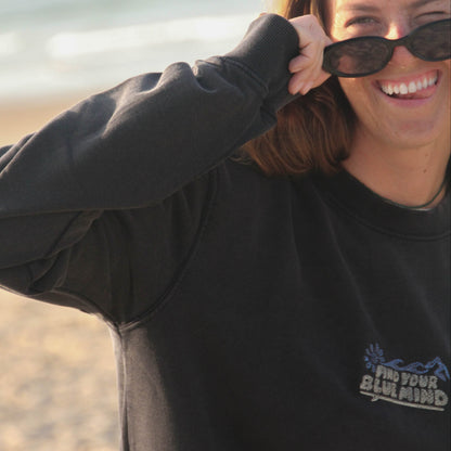 A woman smiling on a beach while wearing a black crewneck sweatshirt with a blue graphic logo that reads 'Find Your Blue Mind' and black sunglasses.
