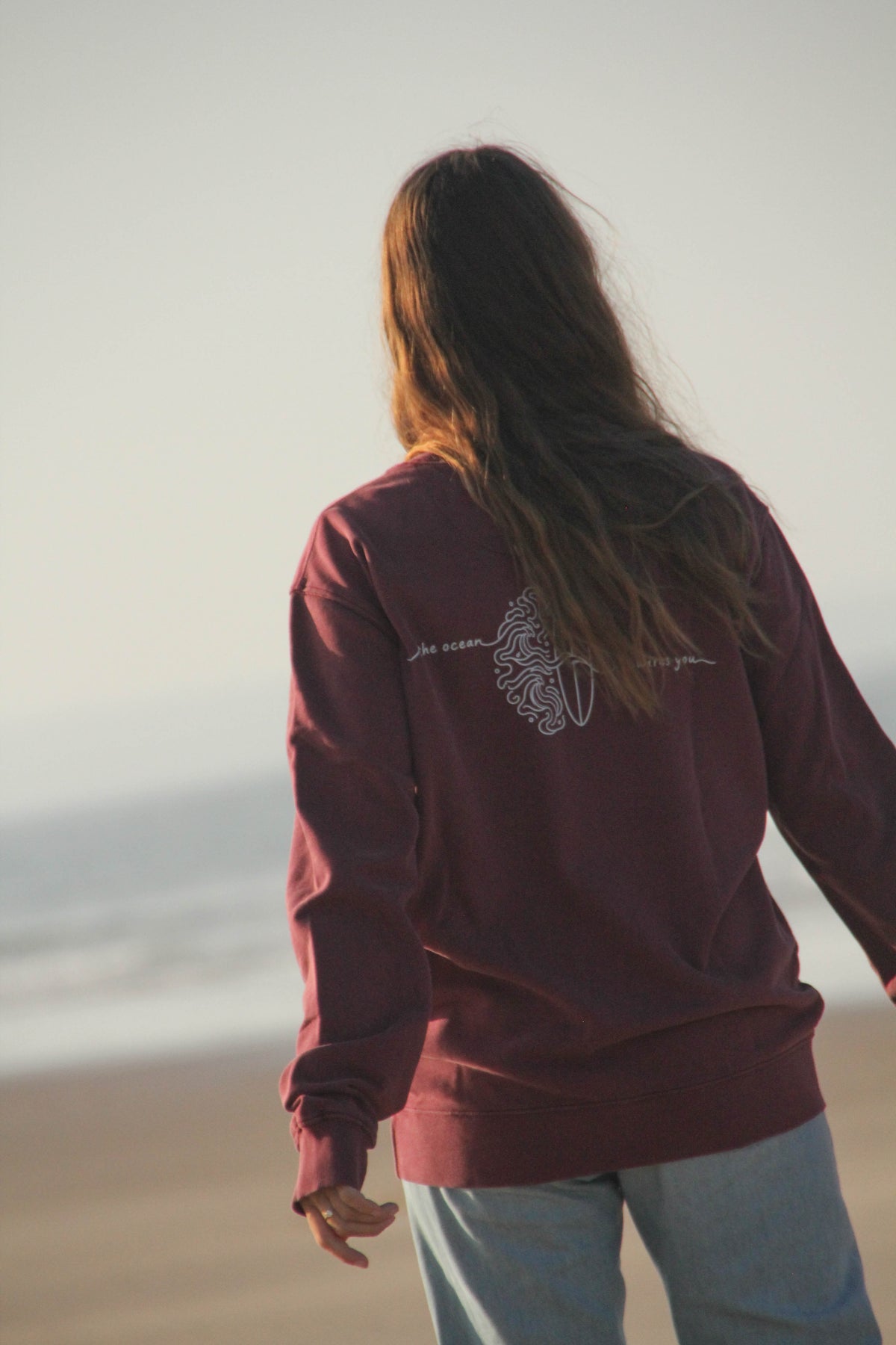 A woman viewed from behind wearing a burgundy crewneck sweatshirt with a white graphic print on the back, standing on a sandy beach.