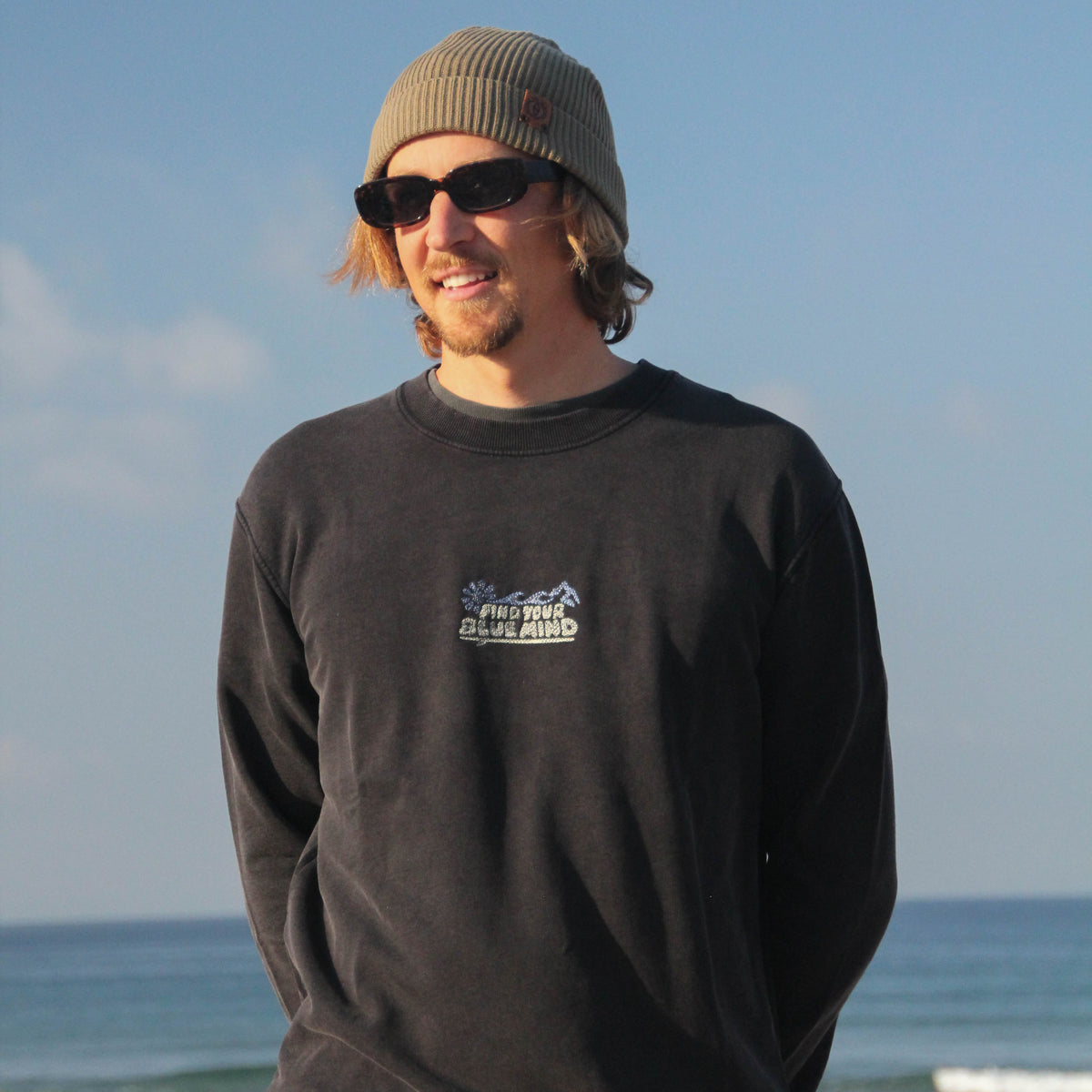 A man wearing a black crewneck sweatshirt with a 'Snow Peak' logo on the chest, a gray beanie, and sunglasses, standing on a beach with the ocean and blue sky in the background.