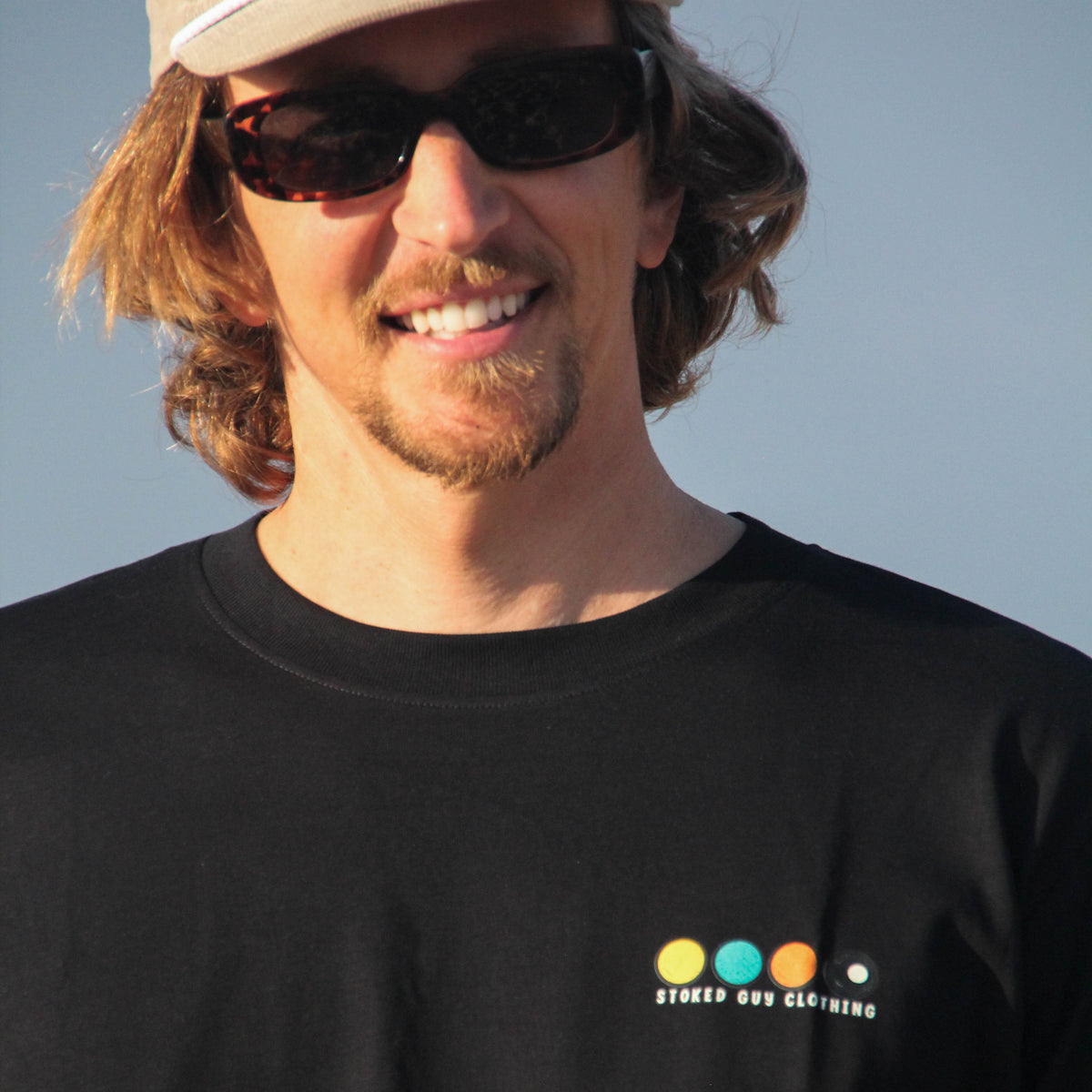 A man wearing a black t-shirt with a colorful logo on the left chest, a beige baseball cap, and sunglasses, photographed outdoors against a clear blue sky.