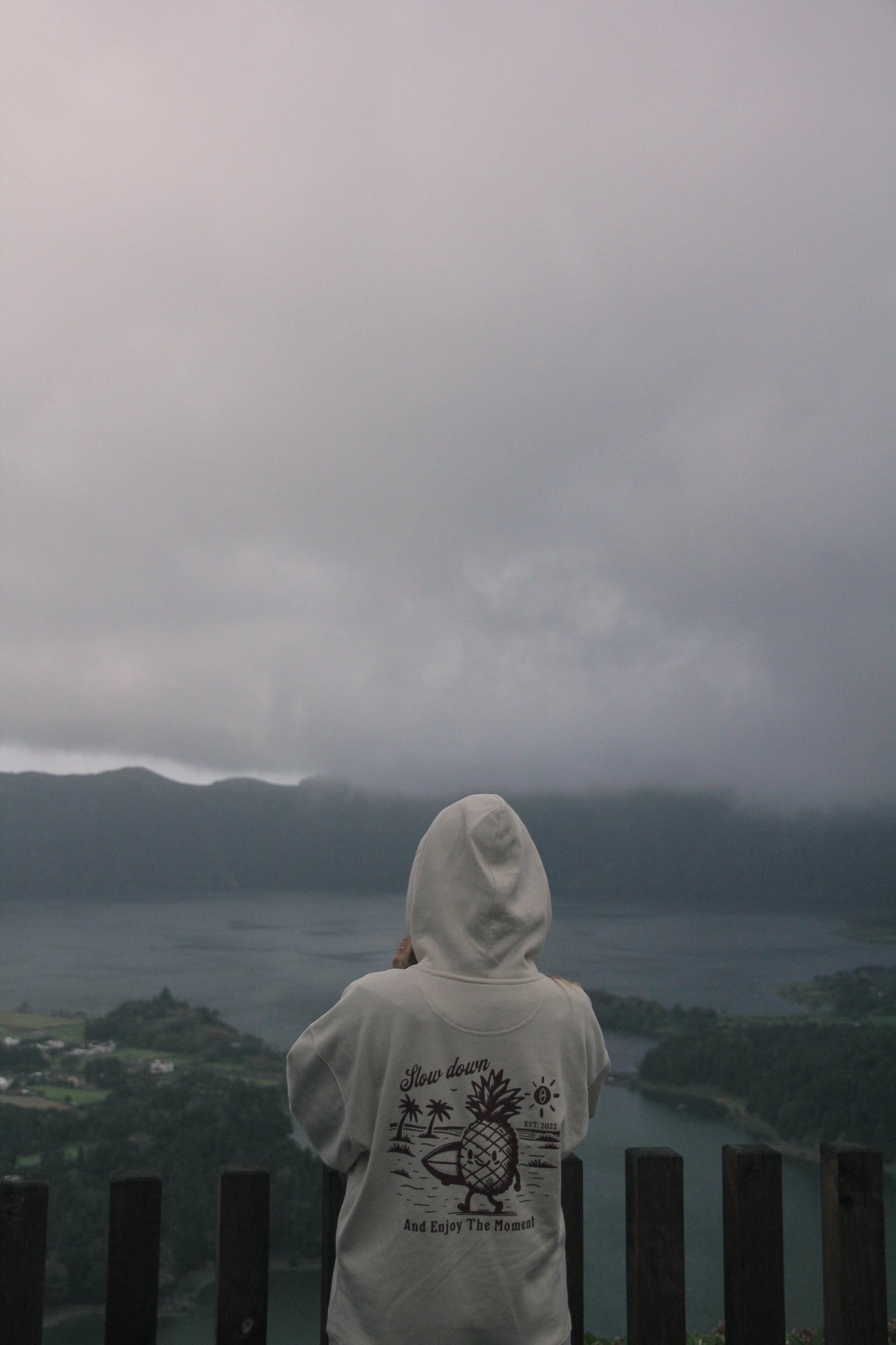 Person wearing a white hoodie with a hood, standing on a wooden platform overlooking a misty landscape.