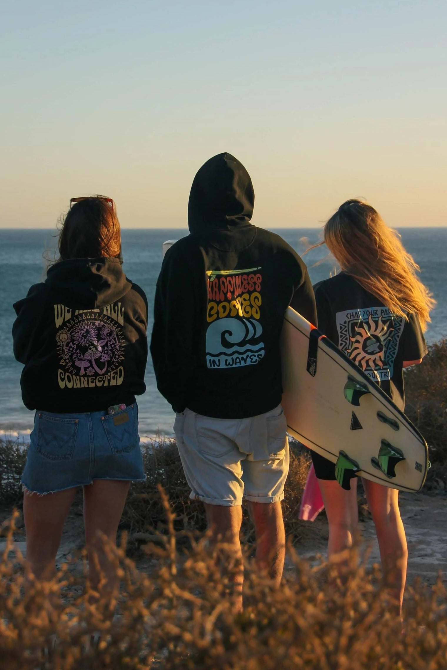 Three people standing on a beach at sunset, wearing hoodies with visible branding.