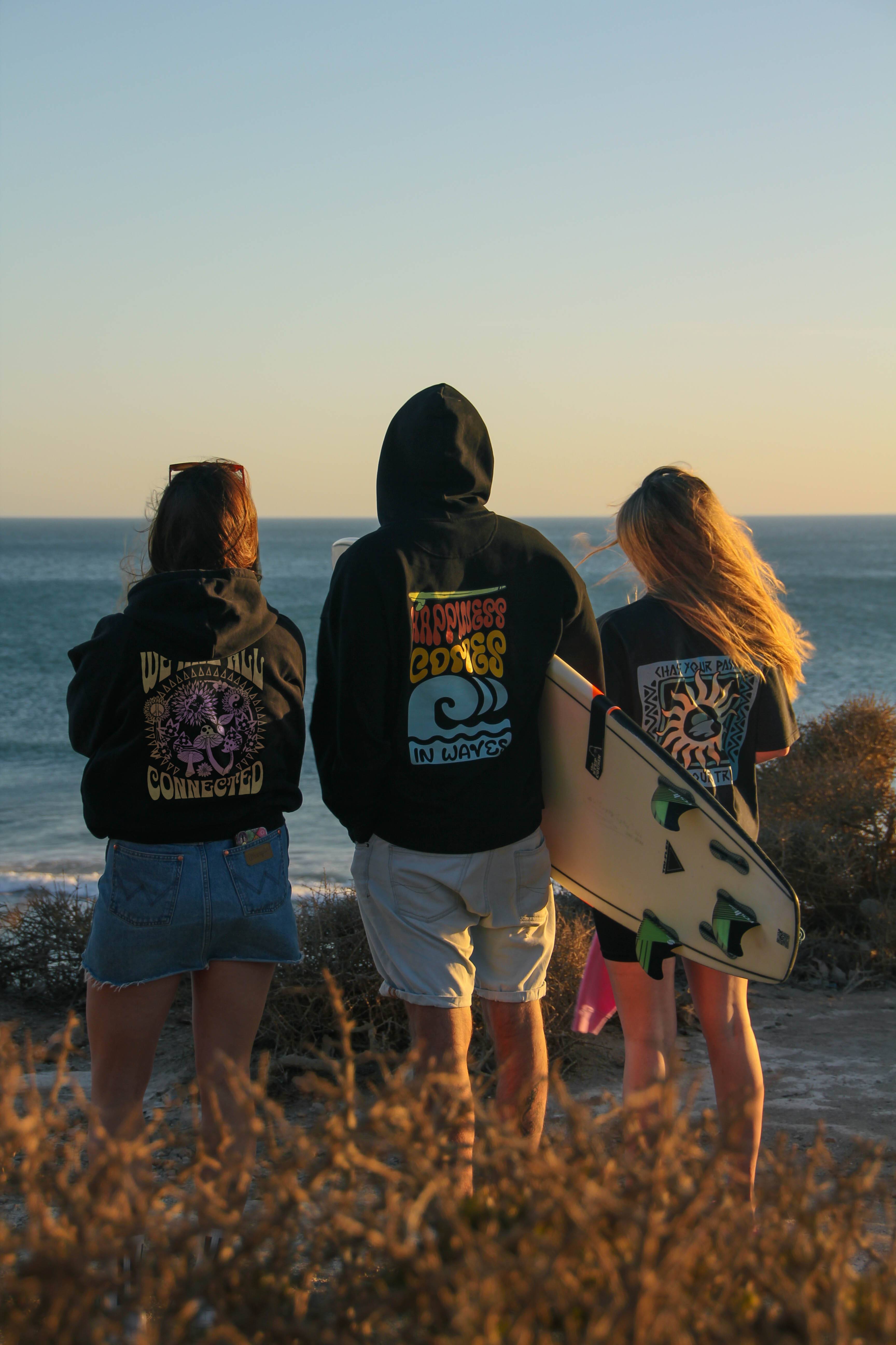 Three people standing on a beach at sunset, wearing hoodies with visible branding.