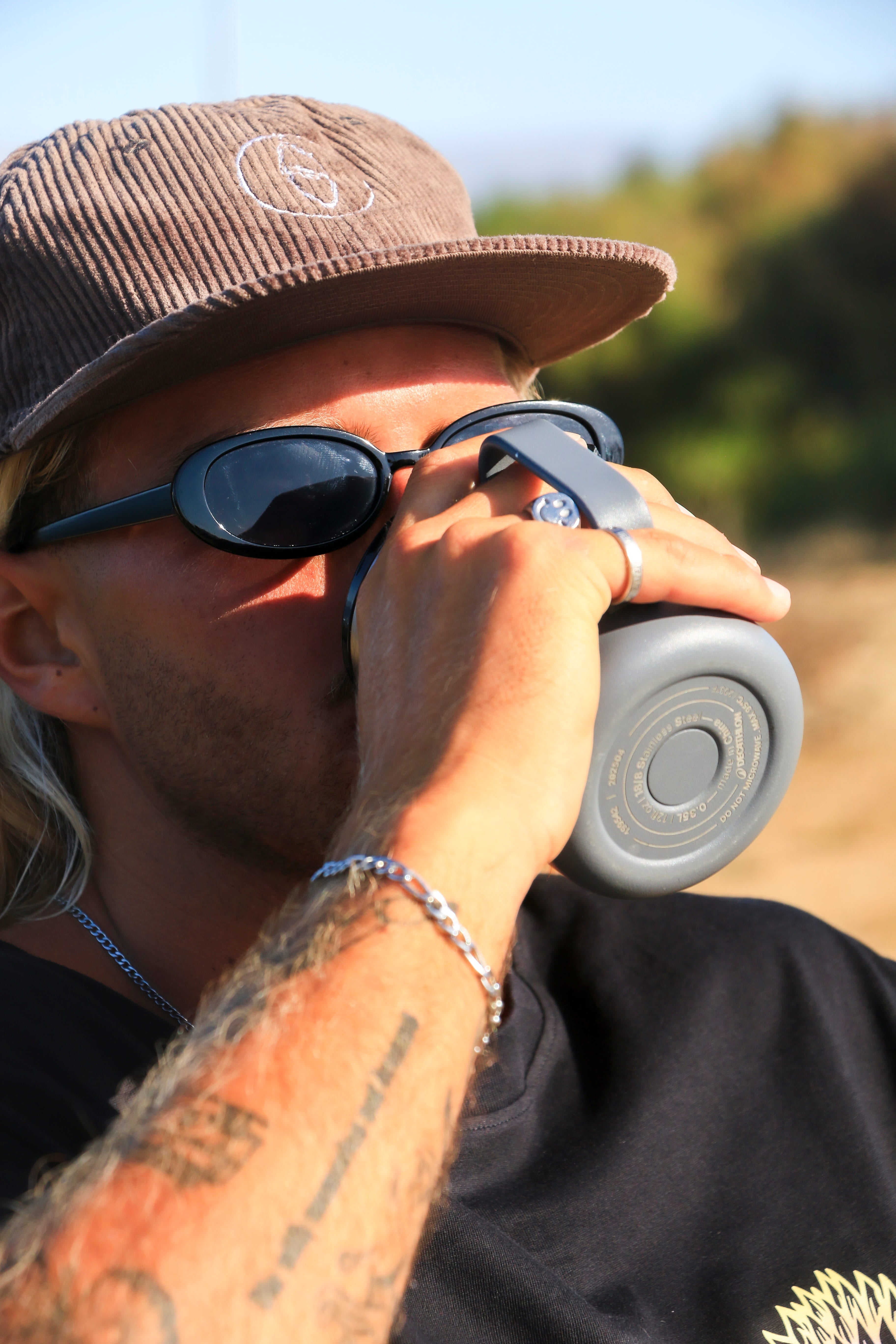 A close-up lifestyle shot of a man wearing a brown corduroy Gucci cap, black sunglasses, and a black t-shirt while drinking from a gray water bottle outdoors.