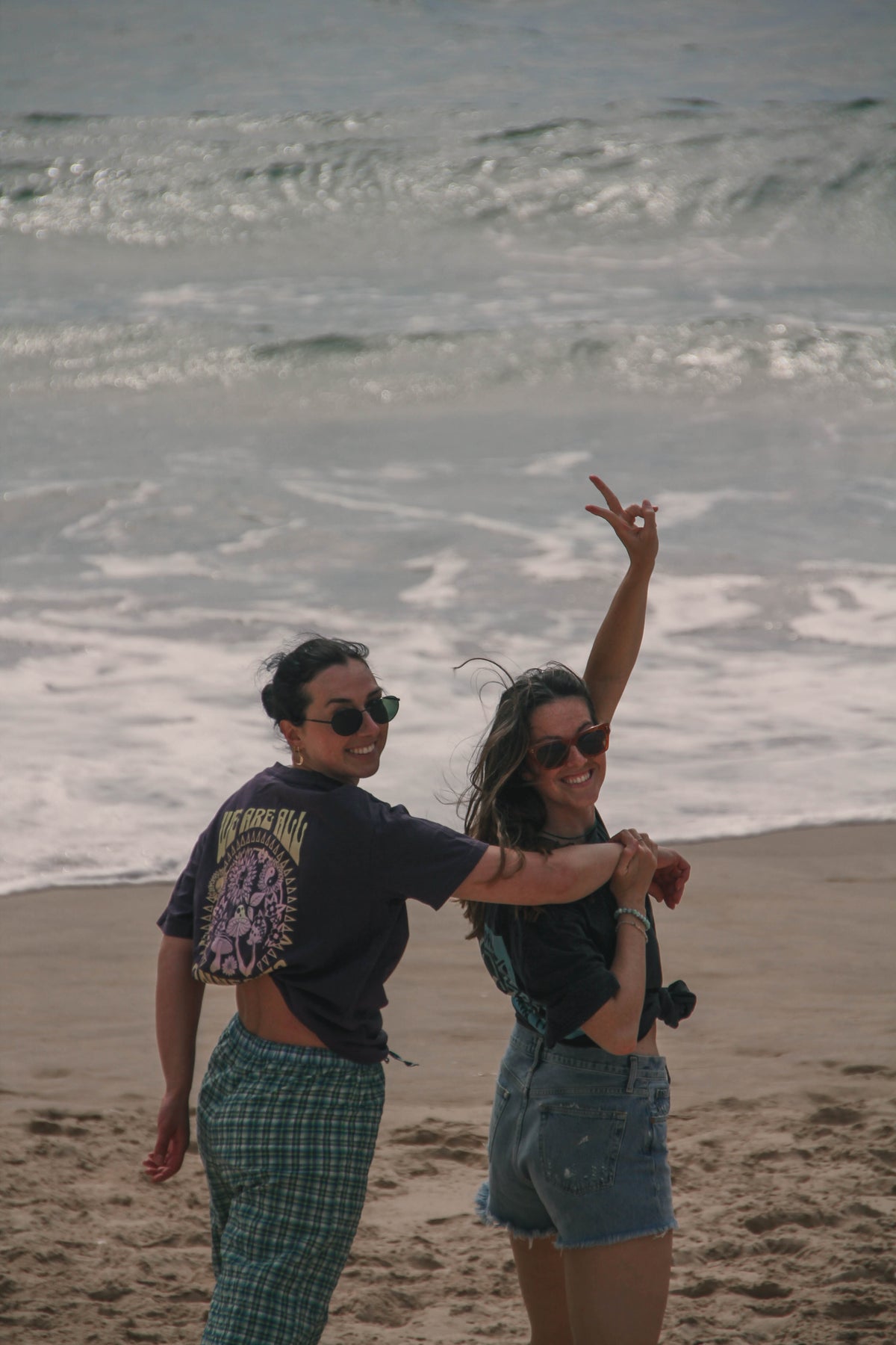 Two women posing on a sandy beach with the ocean waves in the background. The woman on the left wears a dark graphic t-shirt and green plaid pants, while the woman on the right wears a dark crop top and denim shorts with her arm raised in a peace sign.
