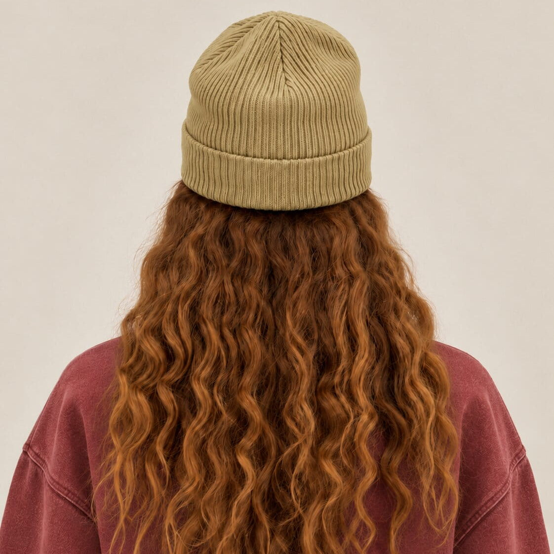 A person shown from the back wearing a ribbed knit beanie in a muted olive-tan color, with long wavy auburn hair and a dusty rose sweatshirt against a plain light beige background.