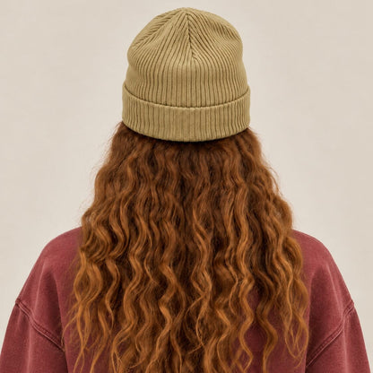 A person shown from the back wearing a ribbed knit beanie in a muted olive-tan color, with long wavy auburn hair and a dusty rose sweatshirt against a plain light beige background.