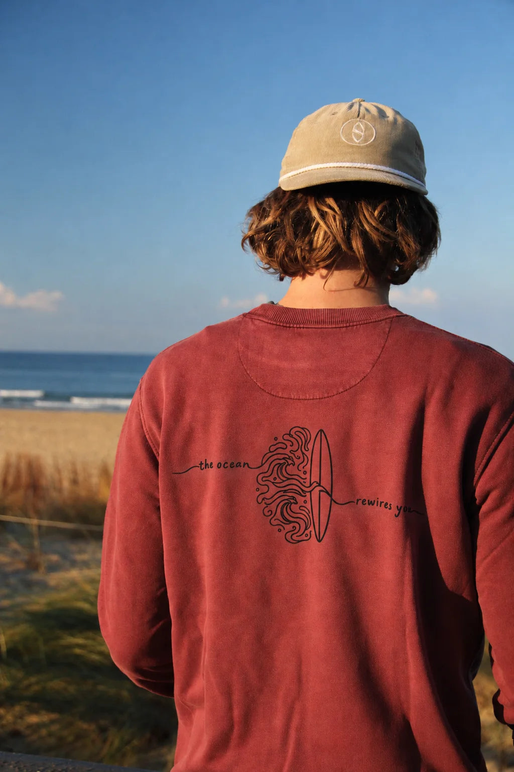 A person viewed from behind wearing a dusty red long-sleeve crewneck sweatshirt and a beige baseball cap, standing on a beach dune with the ocean and sky in the background. The sweatshirt features a black graphic on the back with a surfboard and the words 'the ocean' and 'rewind you'.