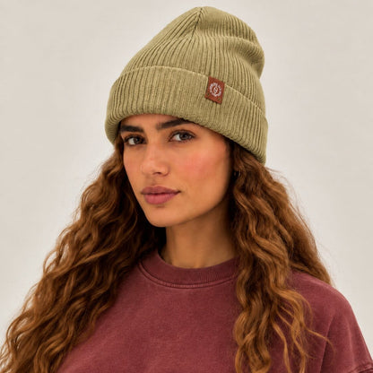 A woman with long wavy light brown hair wearing a ribbed olive green beanie with a small red logo patch and a faded burgundy sweatshirt, photographed against a neutral light gray background.