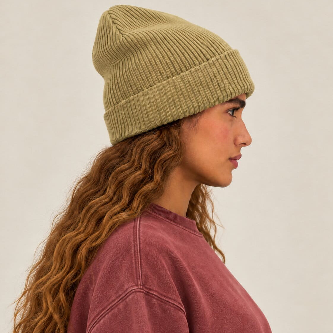 A woman in profile wearing a ribbed knit beanie in a muted olive-khaki color and a dusty rose sweatshirt, photographed against a plain light beige studio background.