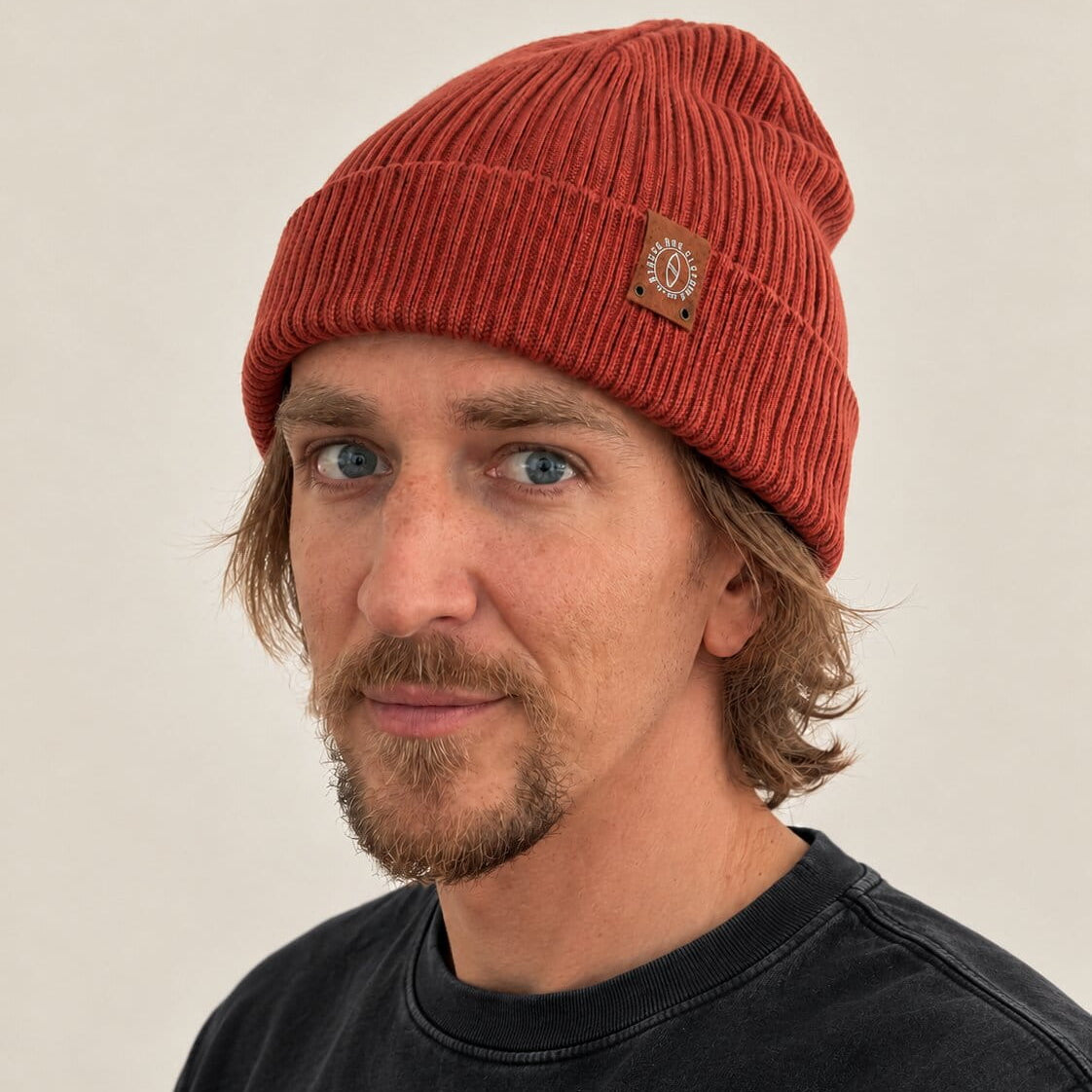 A man wearing a rust-orange ribbed knit beanie with a small brown brand patch on the side, photographed against a plain light beige background.