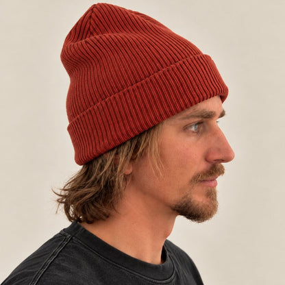 A man in profile wearing a rust-red ribbed knit beanie and a dark charcoal t-shirt, photographed against a plain light beige background.