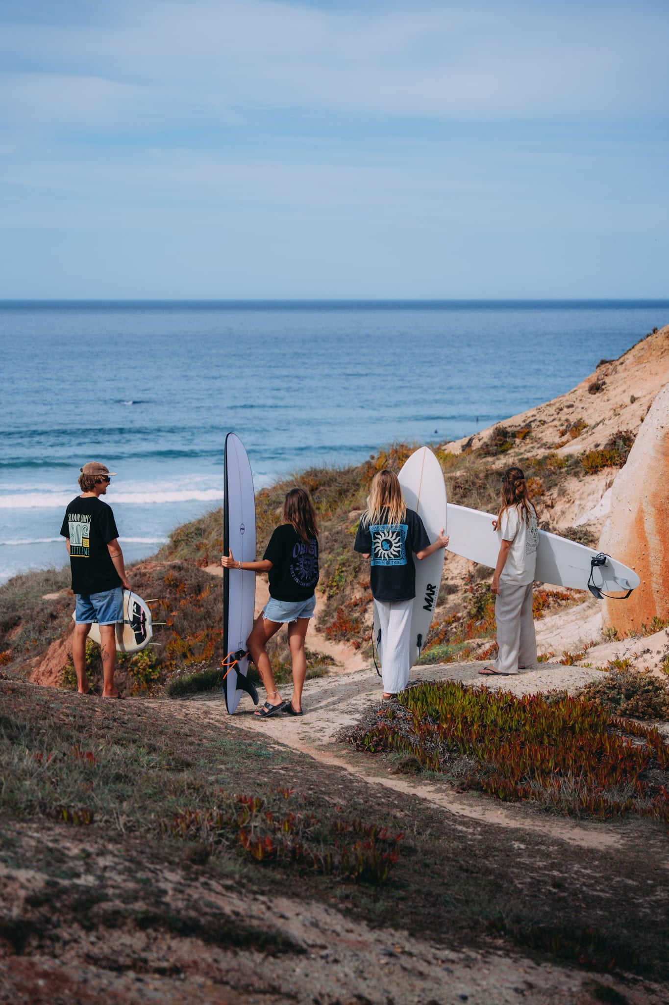 Four people with surfboards walking along a coastal path towards the ocean.