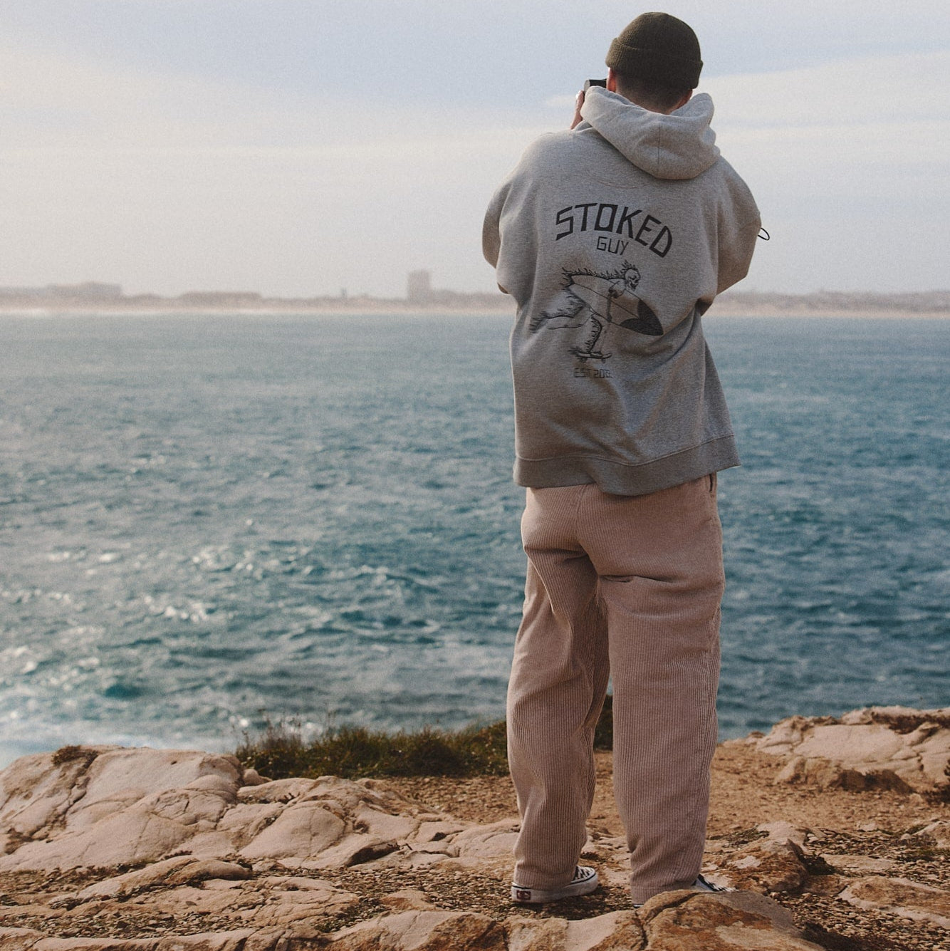 Person wearing a hoodie with 'Stoked Guy' text by the ocean