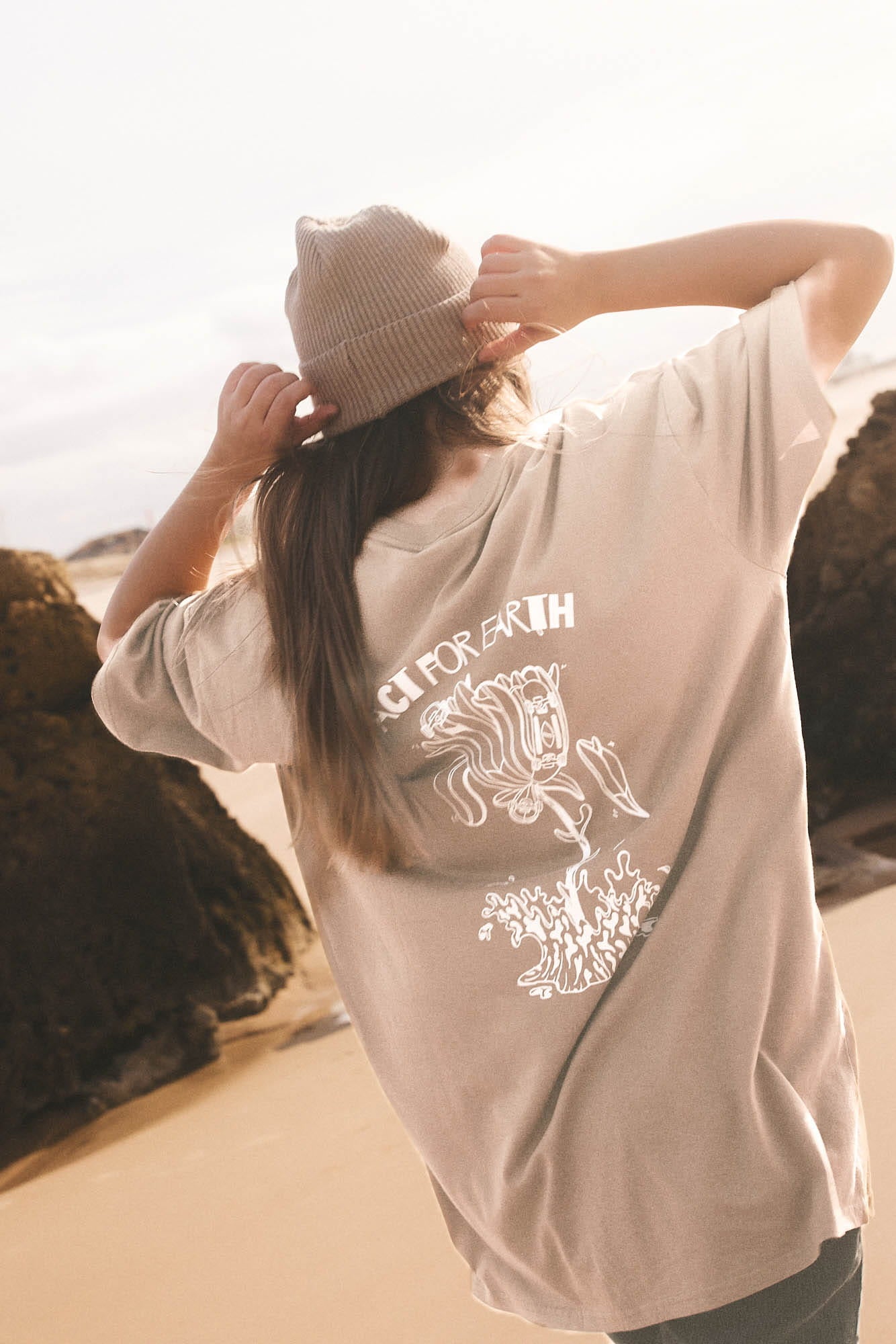 A person standing outdoors on a sandy beach wearing an oversized light beige T-shirt with a white graphic print on the back, a matching beige beanie, and dark pants. The shirt has the words 'CITY FOR GRIEF' printed above a white illustration.