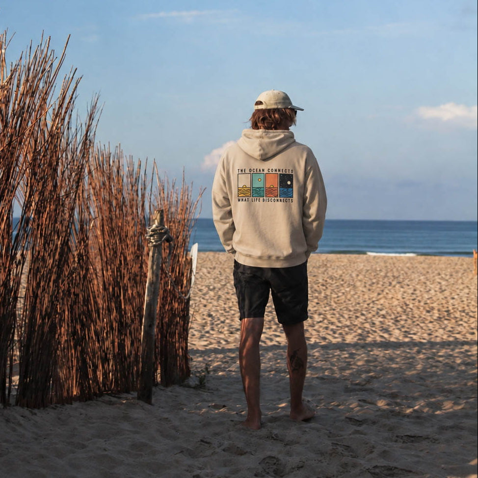A man stands on a sandy beach facing the ocean, wearing a beige hoodie with a colorful graphic on the back, black shorts, and a white cap. Tall dried reeds line the left side of the beach under a blue sky.