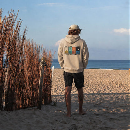 A man stands on a sandy beach facing the ocean, wearing a beige hoodie with a colorful graphic on the back, black shorts, and a white cap. Tall dried reeds line the left side of the beach under a blue sky.