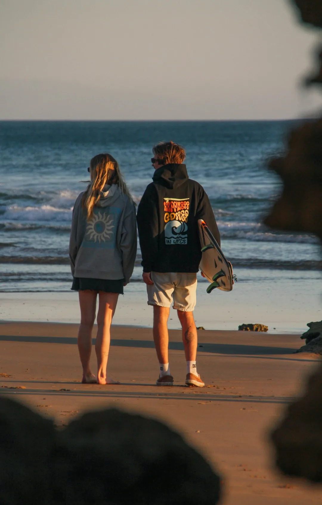 Two people standing on a beach facing the ocean, viewed from behind. The person on the right wears a black graphic hoodie and carries a surfboard, while the person on the left wears a gray hoodie with a sun graphic.