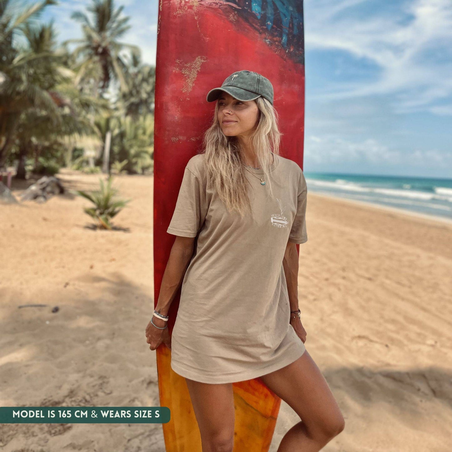 Woman standing on a beach with a surfboard, wearing a beige t-shirt dress and cap.