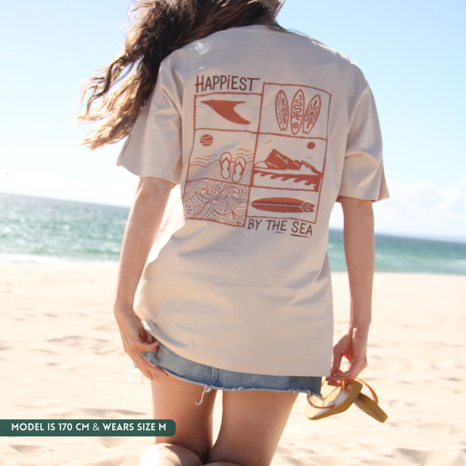 Person wearing a white t-shirt with beach-themed graphics and text on a beach.