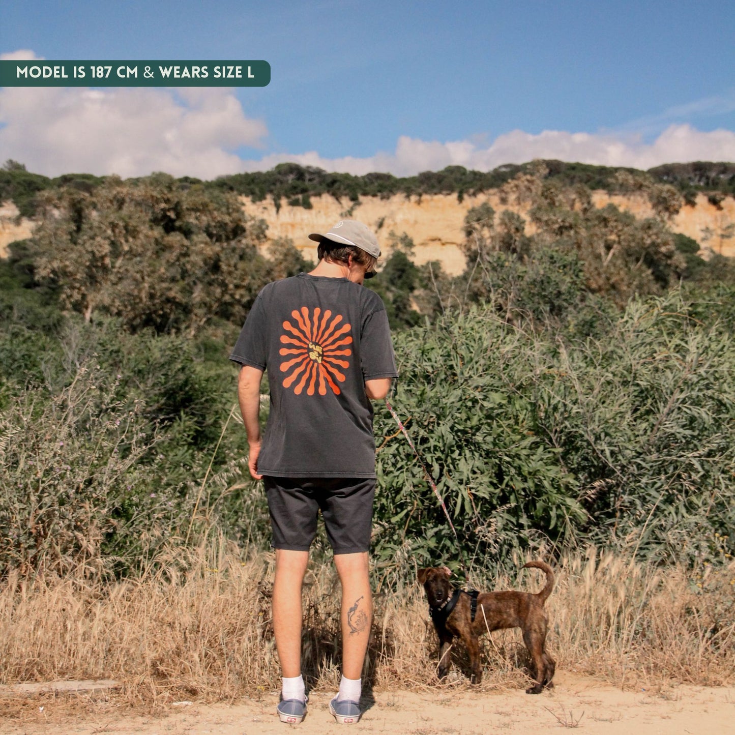 Man walking with a dog in a natural landscape wearing a black t-shirt with a red sun design.