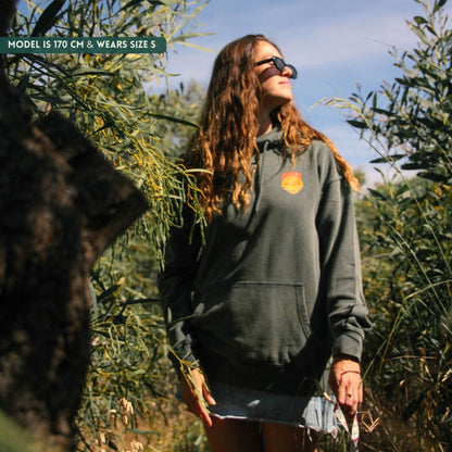 Woman wearing a dark green hoodie with a logo, standing in an outdoor setting with trees.