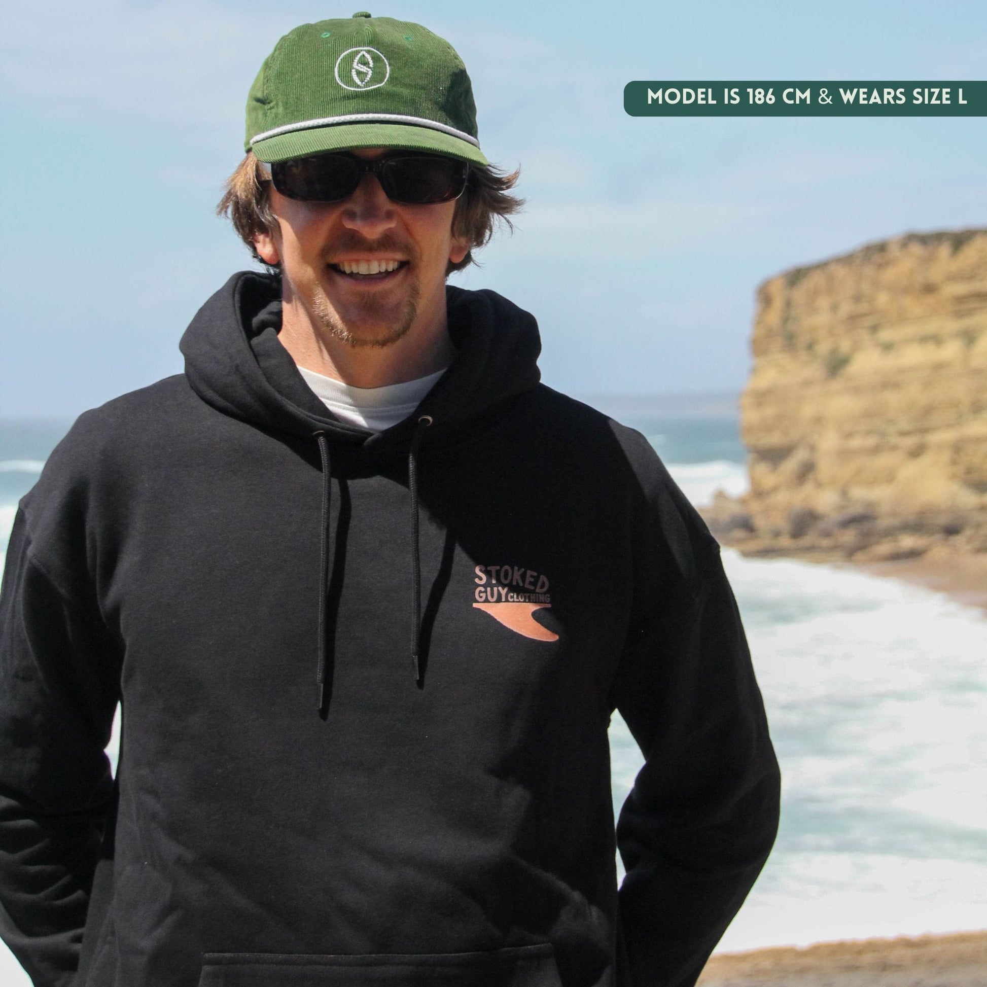 Man wearing a black hoodie with a logo, green cap, and sunglasses on a beach with cliffs in the background.