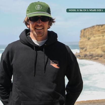 Man wearing a black hoodie with a logo, green cap, and sunglasses on a beach with cliffs in the background.