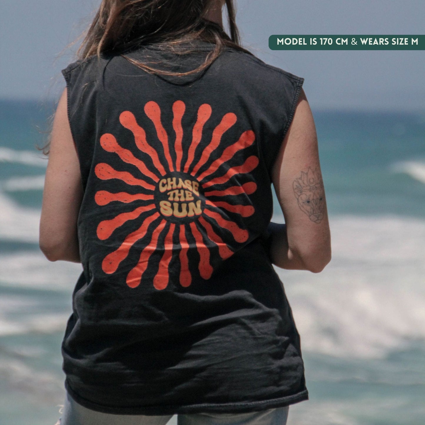 Person wearing a black sleeveless shirt with a red sun design and 'Chase the Sun' text, standing by the ocean.