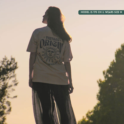 A woman stands outdoors at sunset wearing a cream-colored graphic t-shirt with a black sun design and text on the back, paired with sheer black pants. A text overlay in the top right reads 'MODEL IS 170 CM & WEARS SIZE M'.