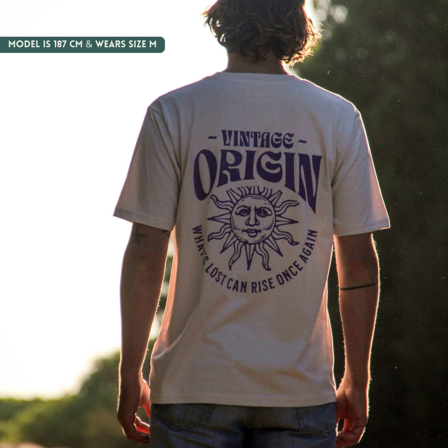 A person viewed from behind wearing a cream-colored t-shirt with a dark purple graphic print on the back. The graphic features the text 'VINTAGE ORIGIN' and 'WHAT'S LOST CAN RISE ONCE AGAIN' surrounding a sun and moon face illustration.