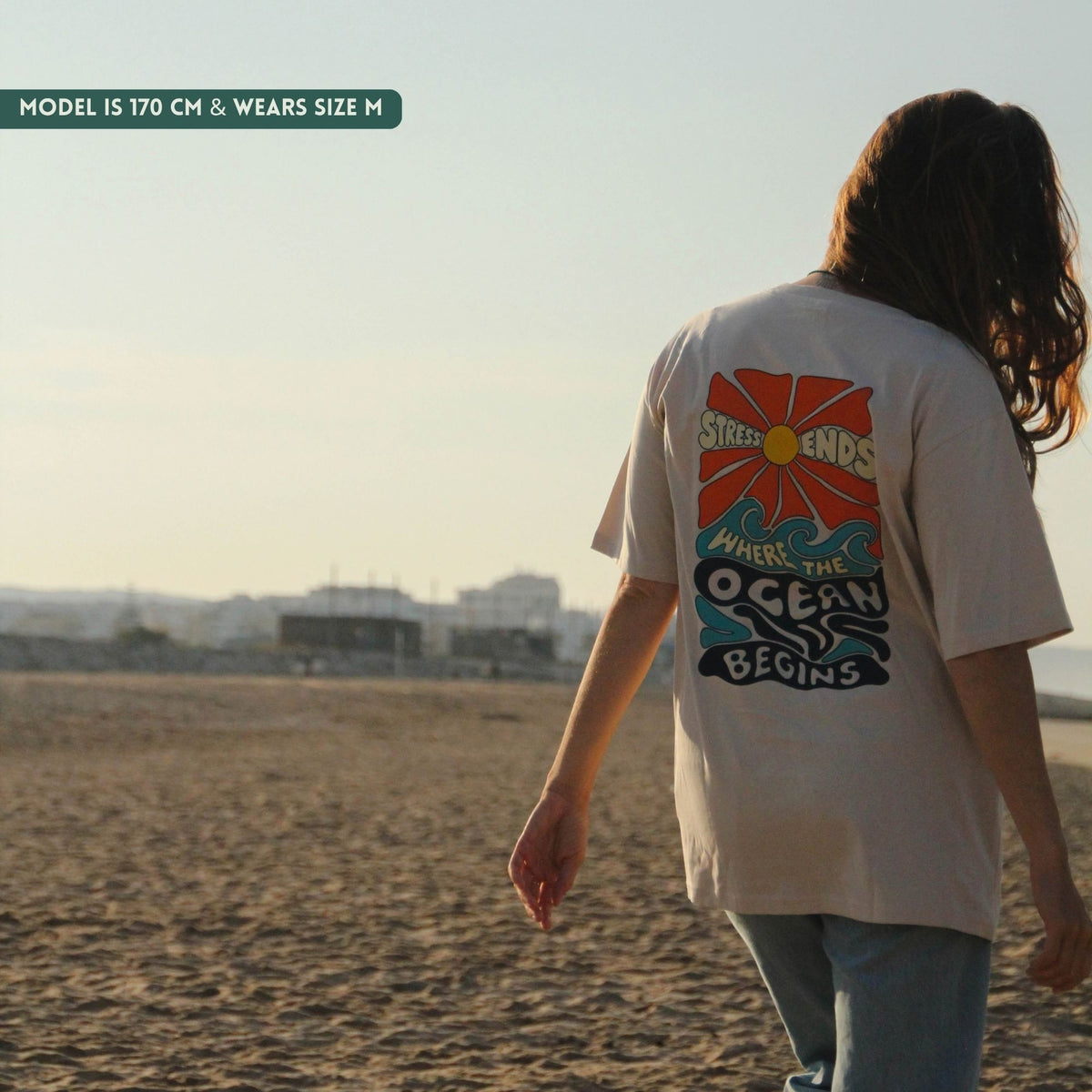 A woman stands on a sandy beach wearing an oversized off-white graphic T-shirt with a colorful sun and ocean design on the back, paired with light blue jeans. The text 'MODEL IS 170 CM & WEARS SIZE M' appears in the top left corner.